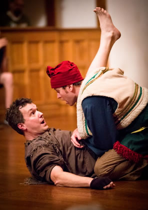 Autylocus in bran ragged clothes on his back as Clown sits astride him, pushing up the rogue's leg: Clown in red stocking cap and fur lined layered peasant clothes